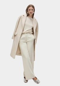 Cappotto in misto lana beige sopra un maglione ecrù, abbinato a pantaloni crema a gamba larga e scarpe basse metalliche argento. Design semplice ed elegante.