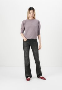 Ung kvinne står med den ene hånden i lommen iført en mauve genser, svarte skinnbukser og røde spisse leiligheter på hvit bakgrunn.