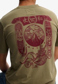 Camiseta verde oliva con un estampado gráfico en marrón de un dragón, una montaña y caracteres asiáticos, con detalles intrincados y acentos circulares.