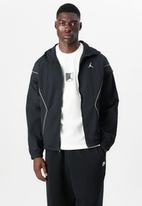 Veste noire avec des accents blancs, en tissu léger, capuche et poignets élastiques. Portée sur un t-shirt blanc avec un logo et un pantalon noir.
