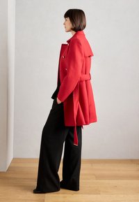 Rode wollen jas met een ceintuur in de taille, dubbelzijdig ontwerp, grote kraag en een gelaagde achterkant, gedragen over zwarte wijde broekspijpen.