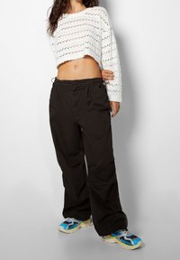 Vit kortärmad tröja med öppet stickmönster, kombinerad med lösa svarta byxor och flerfärgade sneakers med blå, gula och svarta accenter.