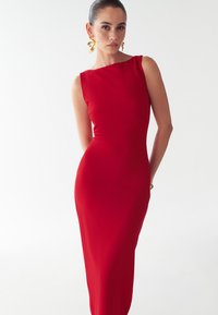 Punane ilma varrukateta kleit, millel on vormitud siluett ja asümmeetriline kaelus, valmistatud siledast kangast. Kuldsed silmapaistvad kõrvarõngad täiendavad välimust.