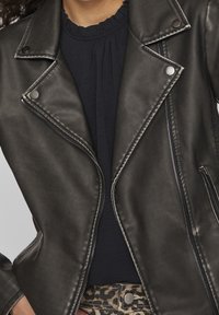 Schwarze Kunstleder-Motorradjacke mit metallischen Nieten, Reißverschluss und strukturiertem Finish, über einem marineblauen Oberteil getragen.