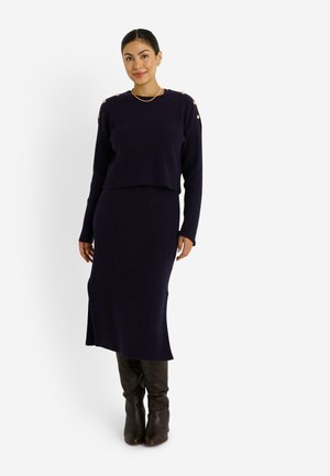 Ensemble en tricot côtelé bleu marine composé d'un pull court et d'une jupe ajustée, avec des accents de boutons dorés aux épaules et une fente sur le côté.