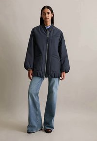 Kvinne med langt mørkt hår iført en svart jakke med glidelås over en blå skjorte og lyseblå flagrende jeans, står foran en ensfarget bakgrunn.