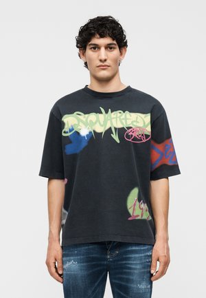 Νέος άνδρας με κοντά σγουρά σκούρα μαλλιά φοράει μαύρο φαρδύ μπλουζάκι με πολύχρωμο κείμενο γκράφιτι και μπλε τζιν σε φθαρμένη όψη, στέκεται μπροστά σε απλό φόντο.