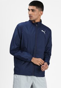 Veste légère bleu marine avec fermeture éclair, manches élastiques et logo blanc sur la poitrine. Texture lisse avec un design minimaliste.