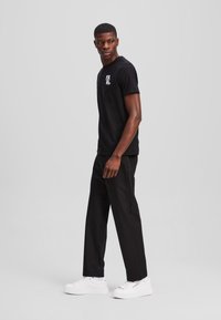 Zwart katoenen t-shirt met logo, gecombineerd met zwarte broeken en witte sneakers. Model staat voor een neutrale achtergrond en toont casual kleding.