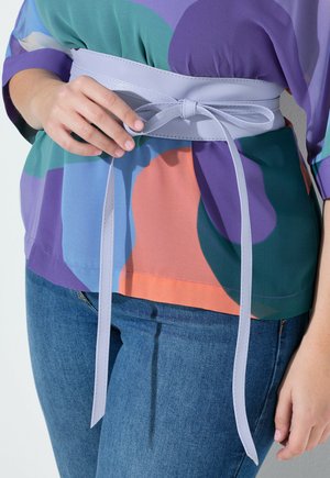 Person trägt blaue Jeans und ein buntes Oberteil in Lila, Orange und Grün und bindet einen breiten, hellvioletten Gürtel um die Taille.