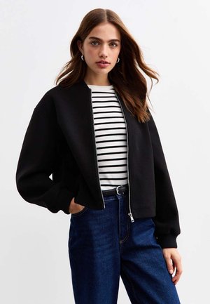 Zwarte cropped bomberjack met een ritssluiting aan de voorkant, ribbels aan de mouwen en een soepele textuur. Gedragen over een gestreept shirt en gecombineerd met blauwe denim jeans.