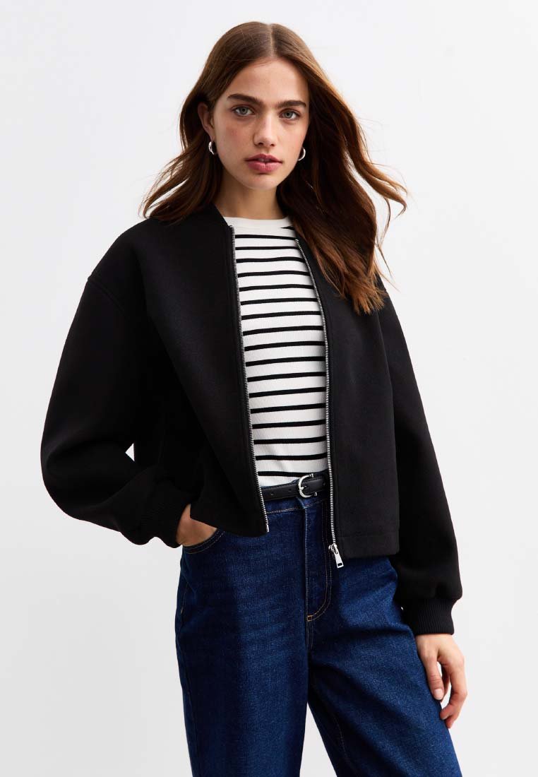 Giacca bomber nera accorciata con zip frontale, polsini a coste e una texture liscia. Indossata sopra una camicia a righe e abbinata a jeans in denim blu.
