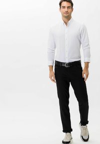 Camicia bianca a maniche lunghe, pantaloni neri slim-fit, cintura nera e sneakers con dettagli. Tessuti uniformemente strutturati, vestibilità su misura e design classico.