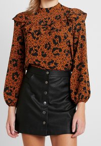 Blusa a maniche corte arancione e nera con motivo a stampa e dettagli arricciati sulle spalle, abbinata a una mini gonna nera in finta pelle con chiusura a bottoni frontale.