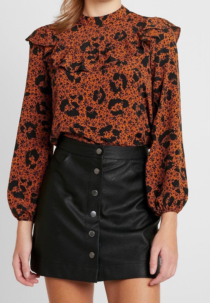 Blusa a maniche corte arancione e nera con motivo a stampa e dettagli arricciati sulle spalle, abbinata a una mini gonna nera in finta pelle con chiusura a bottoni frontale.