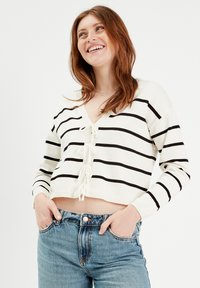 Suéter blanco recortado con rayas negras, escote en V y detalle de lazo en la parte delantera. Combinado con jeans de color azul claro. Textura de punto suave.