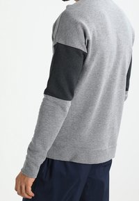 Harmaa huppari, jossa on mustat tekstuuriset hihat, pyöreä kaula-aukko ja resorit. Takakuvassa korostuvat vastakkaiset kankaat ja siistit linjat.