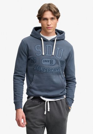 Jonge man draagt een blauwe hoodie met een grote zak aan de voorkant en tekst, gecombineerd met donkergrijze sweatpants met een witte trekkoord.