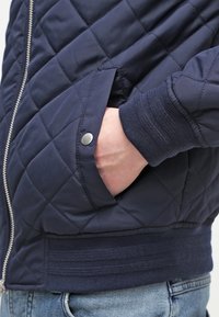 Ręka w kieszeni granatowej pikowanej kurtki z zamkiem błyskawicznym i zatrzaskiem, noszonej z niebieskimi dżinsami.