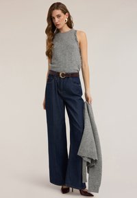 Szary sweter bez rękawów z ozdobami po bokach, zestawiony z szerokimi, ciemnoniebieskimi dżinsami i brązowym paskiem z okrągłą klamrą. Na ramionach narzucony szary kardigan.