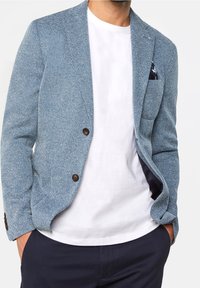 Blå texturerad kavaj med två bruna knappar, en enda bröstficka och en vit t-shirt under, i kombination med mörka byxor.