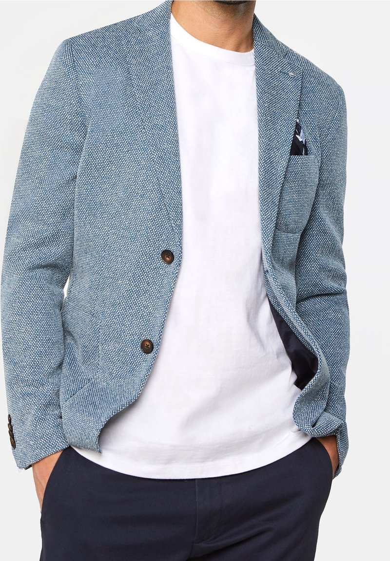 Blå texturerad kavaj med två bruna knappar, en enda bröstficka och en vit t-shirt under, i kombination med mörka byxor.