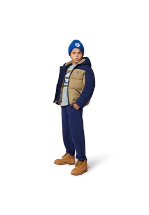 Gevoerde marineblauwe en tan jas met een capuchon, blauwe gebreide beanie, lichtblauw T-shirt met opdruk, marineblauwe broek en tan laarzen met gele veters.