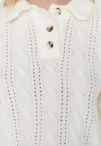 Weißer Strickpullover mit einem Zopfmuster, einem Kragen und drei goldfarbenen Knöpfen an der Knopfleiste. Weiche Textur mit offenen Strickpartien.