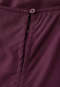 Burgundy stof med en knaplukning for oven og en central søm, med en subtil tekstur fremhævet af plissee-detalje.