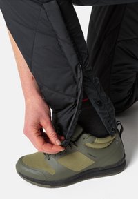 Pantalons isolés noirs avec un design matelassé, présentant une ouverture latérale zippée. La personne ajuste le poignet sur une chaussure verte.