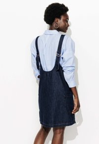 Robe salopette en denim bleu foncé, avec bretelles réglables, une poche et une forme évasée, associée à une chemise rayée bleu clair.