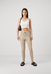 Witte, cropped tanktop met geschulpte randen, beige gestreepte elastische broek en zwarte slide sandalen. Een kleine zwarte schoudertas wordt gedragen.
