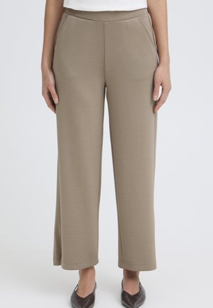 Pantalon beige à jambes larges avec poches latérales porté par une personne avec les mains reposant sur les côtés, associé à des chaussures pointues foncées.