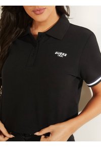 Černé polo tričko s límečkem, krátkými rukávy s bílými a modrými pruhy a bílým logem "GUESS 1981" na hrudi.