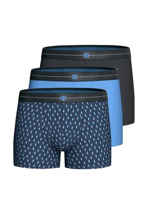 Joven hombre en forma con cabello corto y castaño que lleva puestos calzoncillos bóxer azules con cintura negra sobre un fondo liso.