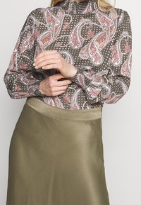 Chemisier à manches longues en vert olive foncé avec un motif paisley rouge et blanc. Présente un col haut froncé et des poignets ajustés. Tissu lisse.