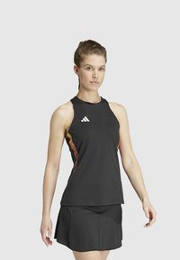 Vrouw in een zwart mouwloos sporttop met oranje zijstrepen en een bijpassende zwarte rok, staande tegen een effen lichte achtergrond.