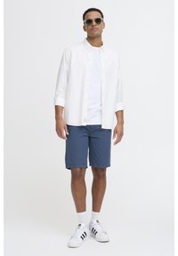 Camisa blanca de botones sobre una camiseta blanca básica, shorts azules y zapatillas blancas con rayas negras. Unas gafas de sol completan el conjunto.