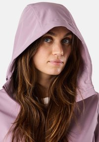 Helle lila wasserdichte Jacke mit Kapuze, die eine glatte Textur und sichtbare Nähte aufweist, getragen über einem weißen Oberteil.