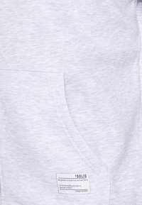 Sudadera gris claro con una textura suave, que presenta un bolsillo frontal y una etiqueta con instrucciones de cuidado. Diseño simple y estética casual.
