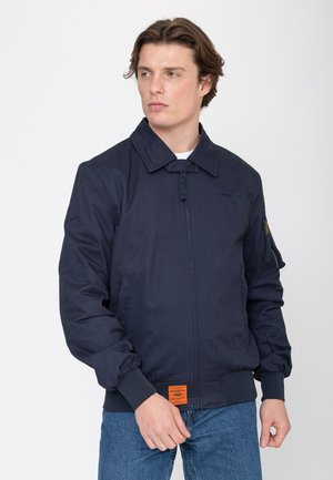 Junger Mann trägt eine marineblaue Bomberjacke mit Frontreißverschluss, gerippten Bündchen und einem orangefarbenen Label nahe dem Saum, kombiniert mit blauen Jeans.