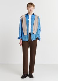Camicia di jeans blu con un maglione beige appoggiato sulle spalle, t-shirt bianca sotto, pantaloni marroni e scarpe scure. Sfondo bianco uniforme.