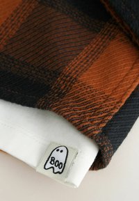 Geweven plaid stof met een oranje en zwarte patroon; witte manchetten met een "Boo" spooklabel. Textuur, zacht materiaal met contrasterende kleuren.