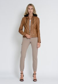 Veste en cuir marron avec capuche noire, fermeture éclair et détails côtelés, associée à un pantalon de jogging beige et des talons noirs à lanières.