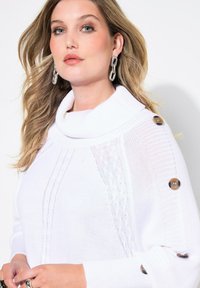 Witte gebreide trui met een col, featuring textuurpatronen en grote bruine knopen op de mouwen. Het model is geaccessoireerd met kettingoorbellen.