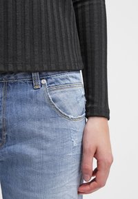 Personne portant un jean en denim bleu clair avec une légère usure et un haut noir à manches longues côtelé, la main détendue le long du corps.