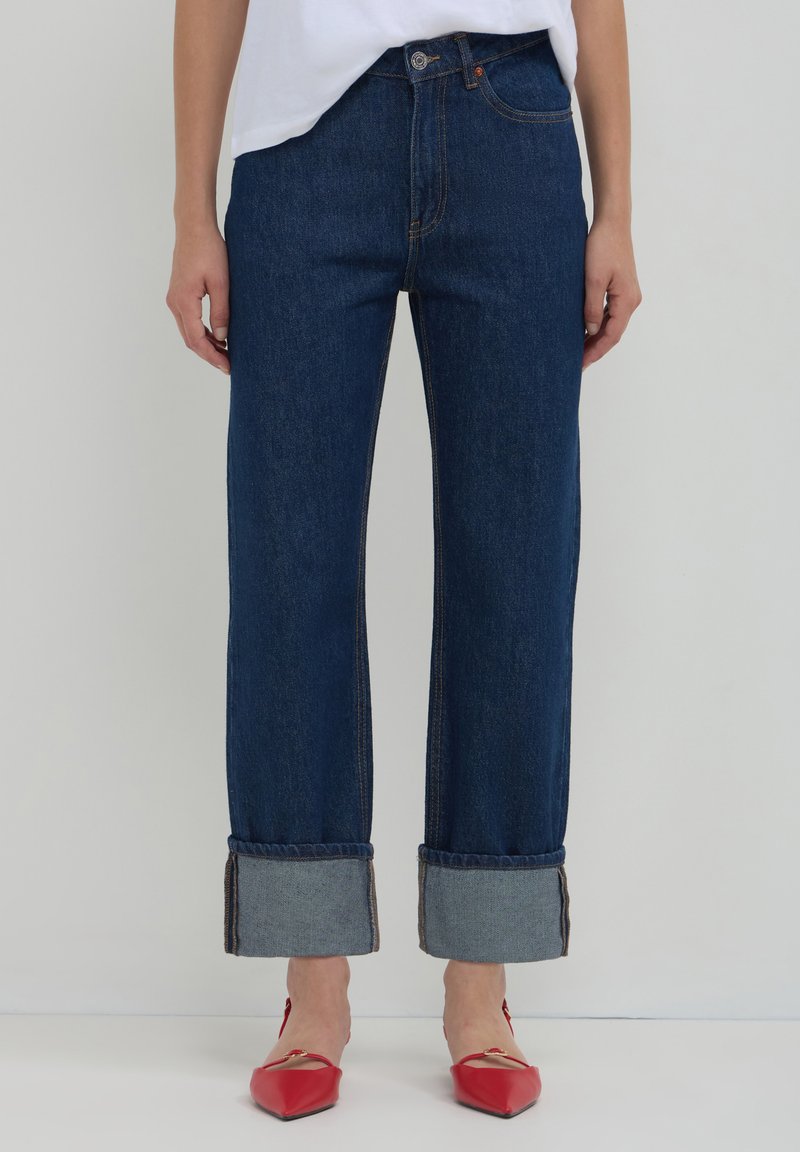 Jeans en denim bleu foncé avec une jambe droite, retroussés au niveau de l'ourlet pour exposer l'intérieur bleu clair, dotés de surpiqûres orange et de boutons à l'avant.