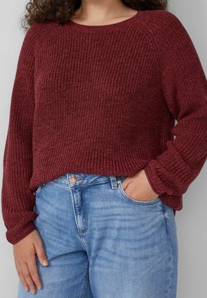 Personne portant un pull en tricot bordeaux et un jean bleu clair, une main dans la poche et l'autre reposant à ses côtés.