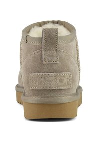 Beige Wildleder-Ankle-Boot mit weichem Futter aus Fell, einer Zuglasche hinten, genähten Details und einer hellbraunen Gummisohle mit Logo-Akzent.