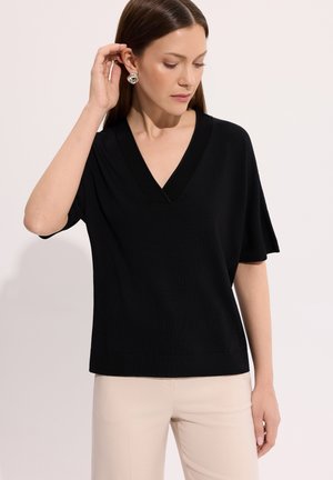 Kobieta poprawiająca srebrną kolczyk w kształcie kwiatu, ubrana w czarny top z krótkim rękawem i dekoltem w serek oraz beżowe spodnie na jednolitym tle.